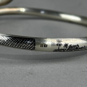 May include: A silver bangle bracelet with etched details. The bracelet features a textured pattern and a landscape scene with palm trees and a hut. The bracelet has a smooth, curved design.