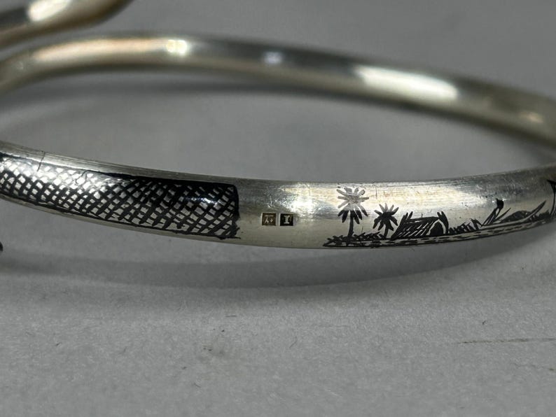 May include: A silver bangle bracelet with etched details. The bracelet features a textured pattern and a landscape scene with palm trees and a hut. The bracelet has a smooth, curved design.