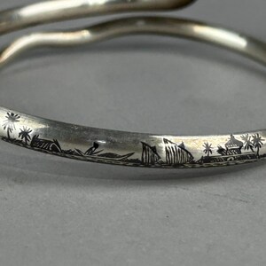 May include: A silver-toned bangle bracelet with an engraved landscape design. The bracelet features a continuous scene of palm trees, buildings, and boats. The bracelet has a smooth, curved design and is shown against a gray background.