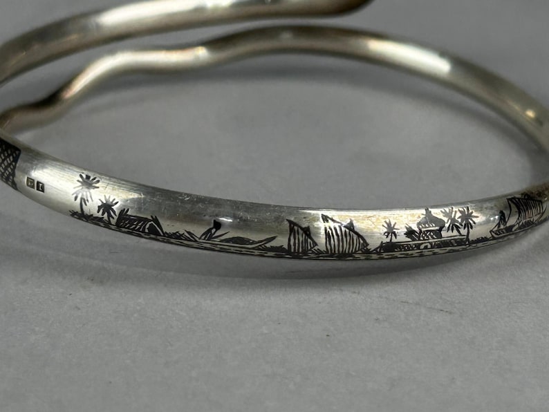 May include: A silver-toned bangle bracelet with an engraved landscape design. The bracelet features a continuous scene of palm trees, buildings, and boats. The bracelet has a smooth, curved design and is shown against a gray background.