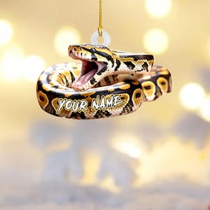 May include: A hanging ornament shaped like a coiled ball python with a brown, black, and yellow patterned scale design. The snake has its mouth open, revealing pink interior details. The text YOUR NAME is printed in white on the snake's body.