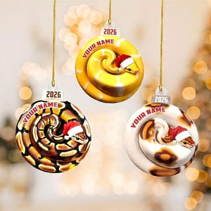 May include: Three circular Christmas ornaments hanging by gold strings. Each features a coiled snake wearing a small red and white Santa hat. The ornaments display the text 2026 and a placeholder for a name in red lettering on the top section.