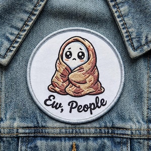 May include: Embroidered patch featuring a cartoon character wrapped in a blanket, with the text "Ew, People." The character has a white face, large eyes, and is covered in a brown blanket. The patch is round with a white border.