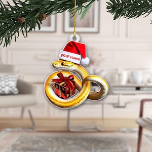 May include: A flat, hanging Christmas ornament featuring a yellow snake coiled around a red gift box with a bow. The snake wears a red and white Santa hat with the text "YOUR NAME" printed on the brim. The ornament hangs from a gold string.