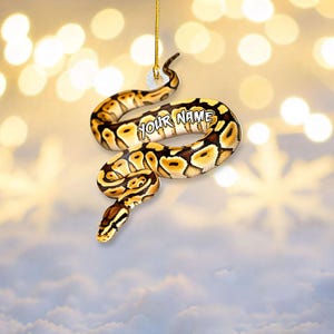 May include: A hanging ornament shaped like a coiled ball python with a brown, black, and yellow patterned skin. The snake features the text "YOUR NAME" printed in white on its body. A gold-colored string is attached to the top for hanging.