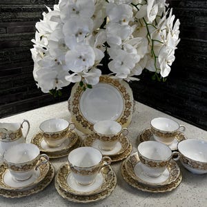 Puede incluir: Un juego de té vintage con tazas, platillos y una cremaera de porcelana blanca, adornado con diseños de hojas doradas. Un gran arreglo floral blanco se encuentra en el fondo. El juego incluye un plato de servir y un cuenco.