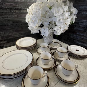 Puede incluir: Un juego de vajilla de porcelana blanca con ribete dorado, que incluye platos, platillos y tazas de té. Un gran ramo de orquídeas blancas en un jarrón blanco está en el fondo.