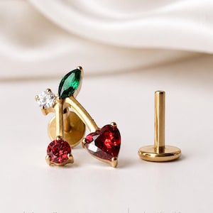 May include: Gold-coloured cherry-shaped earring with red heart-shaped and round gemstones, a green marquise-cut gemstone, and a small white gemstone. The earring is made of hypoallergenic titanium and 18k gold.
