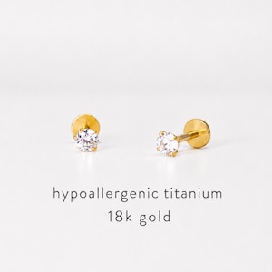May include: Two gold-colored stud earrings with clear, round gemstones. The earrings are set against a white background. Text below reads "hypoallergenic titanium 18k gold."