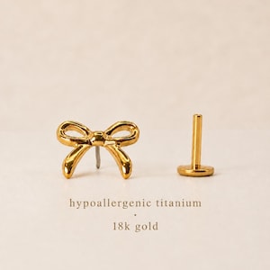 May include: A pair of gold-colored bow-shaped earrings and a gold-colored post. The earrings are made of hypoallergenic titanium and 18k gold. The text "hypoallergenic titanium" and "18k gold" is below the earrings.