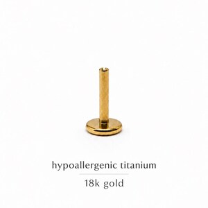Puede incluir: Un pendiente tipo labret de color dorado con parte posterior plana, poste recto y base circular. El texto debajo de la joya dice: hypoallergenic titanium 18k gold. Esta pieza está diseñada para perforaciones corporales y tiene un acabado metálico.