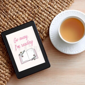 Könnte beinhalten: Ein schwarzer E-Reader zeigt den Text "Go away, I'm reading" über einer Illustration eines offenen Buches mit floralen Akzenten. Eine weiße Teetasse mit Tee steht rechts auf einer Untertasse. Die Artikel liegen auf einer gewebten Matte und einer Holzoberfläche.