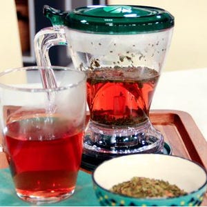 Peut inclure: Un infuseur à thé en verre transparent avec un couvercle et une poignée verts, rempli de thé rouge. Une tasse en verre transparent avec du thé rouge est posée à côté de l'infuseur sur un plateau en bois. Un petit bol de feuilles de thé en vrac est également sur le plateau.