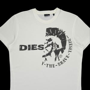 Könnte beinhalten: Cremefarbenes T-Shirt mit Rundhalsausschnitt und großem schwarzem Grafikdruck. Der Druck zeigt das Wort "DIES" und ein stilisiertes Gesicht mit Irokesenschnitt, umgeben von den Worten "DIESEL" und "Y-THE-BRAVE".