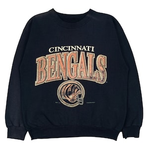 May include: A black crewneck sweatshirt featuring the text CINCINNATI BENGALS in a large, arched, distressed orange and white font. Below the text is a circular graphic of a football helmet with tiger stripes in orange, black, and white.