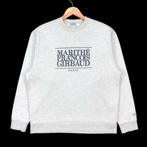 May include: A light grey sweatshirt featuring the navy blue text "MARITHÉ FRANÇOIS GIRBAUD PARIS". The sweatshirt has a crew neck, long sleeves, and ribbed cuffs and hem. It is displayed on a wooden hanger.