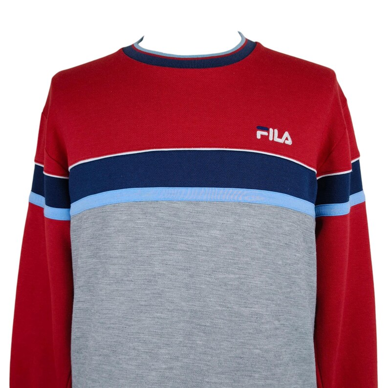 Puede incluir: Una sudadera Fila roja, gris y azul. La parte superior es roja, con una franja azul marino y azul claro en el pecho. La mitad inferior es gris. El logotipo de Fila est&aacute; bordado en la parte superior derecha del pecho.
