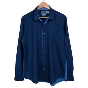 Può includere: Camicia di jeans blu scuro a maniche lunghe, con bottoni e una tasca sul petto. Il colletto e i polsini sono di un blu più chiaro. La camicia è appesa a una gruccia di legno su sfondo bianco.