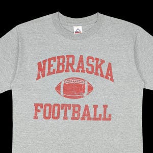 May include: A grey long-sleeved t-shirt featuring the word "NEBRASKA" in red, arched across the top. Below is a red football graphic, and the word "FOOTBALL" is printed in red below.