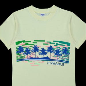 Puede incluir: Camiseta amarillo claro con un diseño tropical. El gráfico incluye palmeras azules, líneas horizontales rosas y verdes, y la palabra "HAWAII" en azul. La camiseta tiene cuello redondo y mangas cortas.