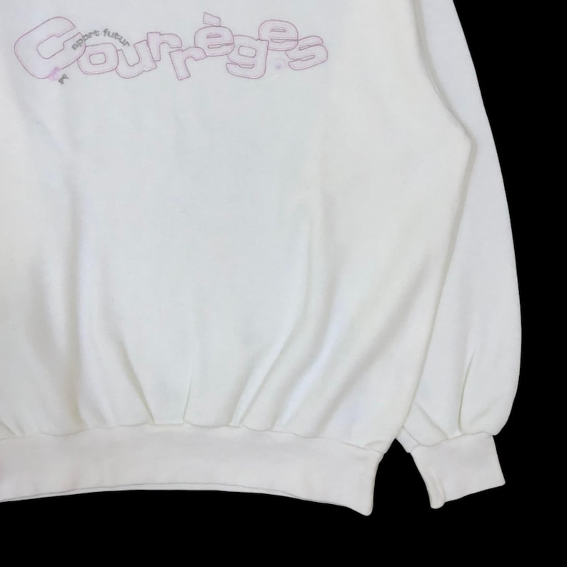 以下が含まれることがあります： 胸元にライトピンクの輪郭線フォントで「Courr&egrave;ges」および「sport futur」の文字がデザインされた、白い長袖のスウェットシャツ。この衣類には、リブ編みのクルーネック、袖口のリブ、裾のリブがあしらわれています。