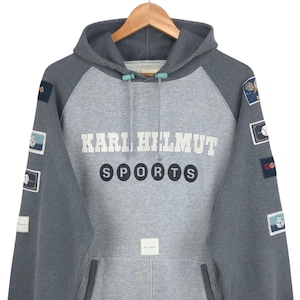 Pode incluir: Um moletom cinza e cinza claro Karl Helmut Sports com capuz com cordão e vários patches nas mangas. A frente apresenta o texto "KARL HILMUT SPORTS" em branco e preto.