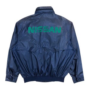 Puede incluir: Chaqueta de nailon azul marino con la palabra "NISSAN" en letras verdes en la espalda. La chaqueta tiene cuello alto, cierre de cremallera y puños y cintura elásticos.