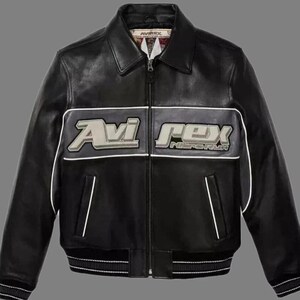 May include: Black leather jacket with the "Avi Rex" logo in white and gray across the chest. The jacket has a ribbed hem and cuffs, a zipper closure, and white trim. A classic bomber jacket style.