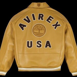 May include: Mustard yellow leather jacket with the word "AVIREX" arched across the back in brown block letters. Below the brand name is a circular emblem and the word "USA". The jacket has a collared neckline and ribbed cuffs and hem.