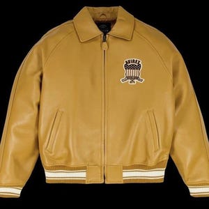 May include: Mustard yellow leather jacket with a collar, zip closure, and ribbed cuffs and hem. Features a chest patch with "AVIREX" and a shield design. The jacket has two front pockets.