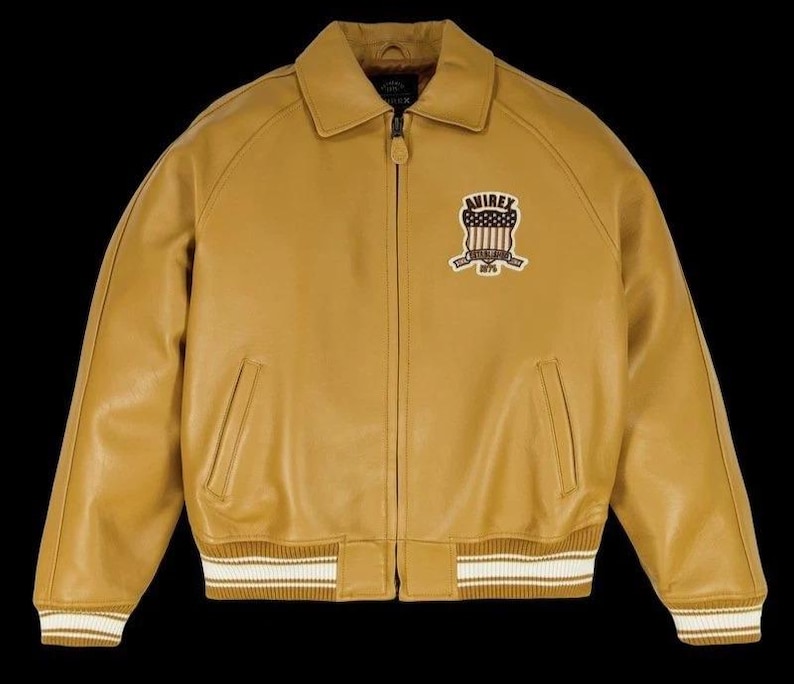 May include: Mustard yellow leather jacket with a collar, zip closure, and ribbed cuffs and hem. Features a chest patch with "AVIREX" and a shield design. The jacket has two front pockets.