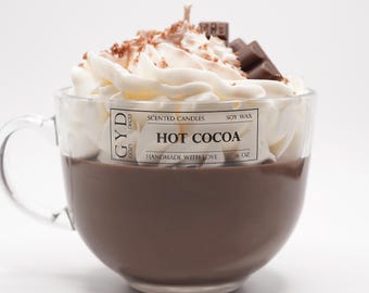 Puede incluir: Una vela con forma de taza de cristal transparente rellena de cera marrón, cubierta con remolinos de cera blanca, cacao en polvo y trozos de chocolate. La etiqueta dice "HOT COCOA". La vela es de 454 gramos.