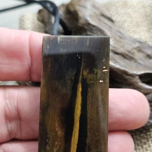 Op de afbeelding: Een rechthoekige hanger met een zwart koord. De hanger heeft een donkerbruine kleur met een verticale gele streep en een houtnerfpatroon. De hanger wordt in de hand van een persoon gehouden.