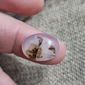 Può includere: Una gemma trasparente di forma ovale con inclusioni marroni e beige. La pietra ha una tonalità viola chiaro traslucida ed è tenuta tra le dita. La gemma è probabilmente un cabochon.