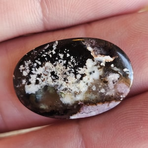 Può includere: Una gemma ovale con una superficie lucida. La pietra presenta una base scura e lucida con inclusioni bianche, beige e viola chiaro. La gemma è tenuta in mano, mostrando le sue dimensioni e la sua consistenza.