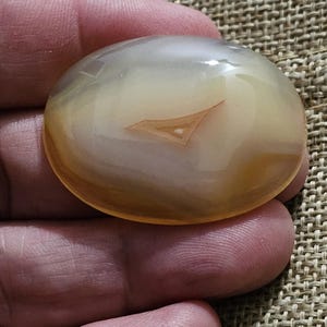 Könnte beinhalten: Ein glatter, ovaler Achat-Stein mit einem weißen und orangefarbenen Bandmuster. Der Stein ist durchscheinend und hat einen leichten Glanz.