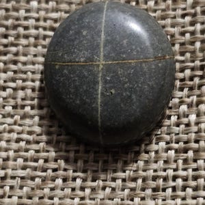 Könnte beinhalten: Ein runder, dunkelgrauer Stein mit zwei weißen Linien, die ein Kreuz bilden.