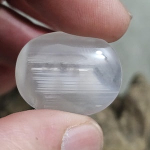 Puede incluir: Una piedra clara y redondeada con inclusiones lineales blancas internas. La piedra se sostiene entre dos dedos, mostrando su calidad translúcida y su textura natural. La forma de la piedra es suave y pulida, reflejando la luz.