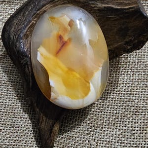 Könnte beinhalten: Ein glatter, ovaler Achat mit einem weißen und orangefarbenen Muster. Der Stein liegt auf einem Stück dunklem Holz.