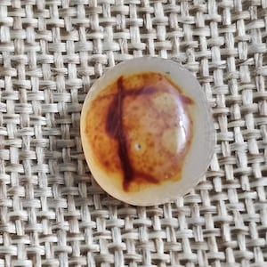 Può includere: Una pietra rotonda e traslucida di colore chiaro con un motivo marrone a forma di croce al centro. La pietra è posta su uno sfondo strutturato e tessuto, creando un'estetica naturale.