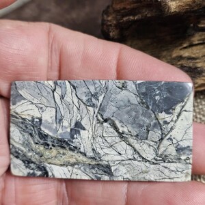 Può includere: Una pietra rettangolare con un motivo marmorizzato nei toni del grigio, nero e crema. La pietra ha una superficie lucida e presenta venature intricate. L'effetto complessivo ricorda un paesaggio naturale.