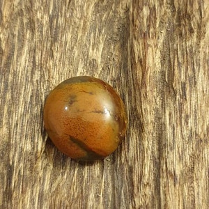 Könnte beinhalten: Ein runder, polierter, brauner und orangefarbener Achat-Stein mit einem wirbelnden Muster. Der Stein liegt auf einem Stück Holz.