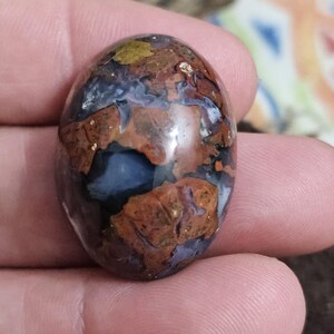 Könnte beinhalten: Ein ovaler Edelstein mit polierter Oberfläche. Der Stein zeigt eine Mischung aus braunen, blauen und goldenen Farbtönen, wodurch ein Marmoreffekt entsteht. Der Edelstein wird in einer Hand gehalten.