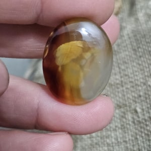 Può includere: Pietra preziosa ovale lucidata con un mix di aree marroni, ambrate e traslucide. La pietra ha una superficie liscia e lucida. I colori e i motivi creano un'estetica naturale unica. La pietra sembra essere tenuta in una mano.