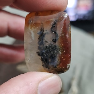 Può includere: Una gemma rettangolare lucidata con una base traslucida, con motivi interni di inclusioni nere e marroni. La pietra ha bordi arrotondati e una finitura lucida, che mostra formazioni minerali naturali.