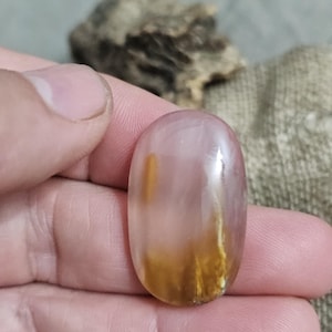 Può includere: Una gemma lucida di forma ovale con una tonalità rosa traslucida. La pietra presenta striature di inclusioni marrone dorato, creando un motivo unico. La gemma è tenuta in una mano, con uno sfondo sfocato.