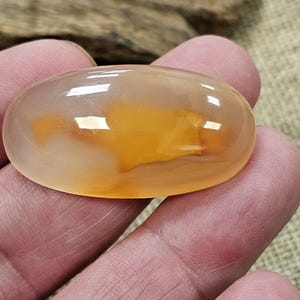 Könnte beinhalten: Ein glatter, ovaler Achat-Cabochon mit einem durchscheinenden, hellorangen und weißen Muster. Der Achat ist poliert und hat eine glänzende Oberfläche.