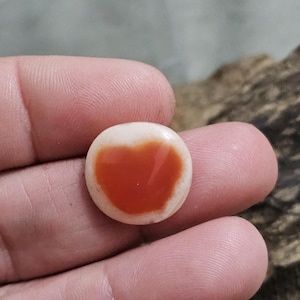 Può includere: Una gemma rotonda e lucida con un bordo bianco crema e un vivace centro a forma di cuore rosso-arancio. La pietra è tenuta tra le dita, mettendo in risalto la sua superficie liscia e riflettente. Il design della gemma ricorda un cuore.