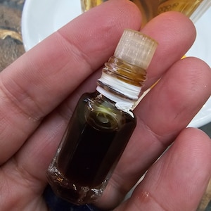 Puede incluir: Una pequeña botella rectangular de vidrio llena de un líquido oscuro, sostenida en una mano. La botella tiene un tapón transparente texturizado y una etiqueta blanca alrededor del cuello. Otras botellas son visibles en el fondo.