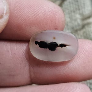 Könnte beinhalten: Ein durchscheinender, oval geformter Edelstein mit einem dunklen, wolkenartigen Einschluss. Der Stein wird zwischen den Fingern gehalten, wodurch seine polierte Oberfläche und seine natürlichen Muster zur Geltung kommen. Die Farbe ist ein blasses, milchiges Weiß mit einem schwarzen Inneren.